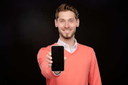 Portrait of sales manager in orange sweater presenting new smartphone and showing it at camera, black backgroundの写真素材