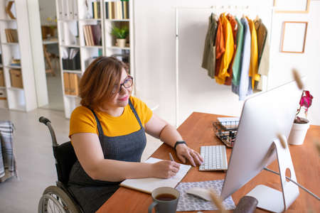 Young disable student in wheelchair making notes in front of computer at home