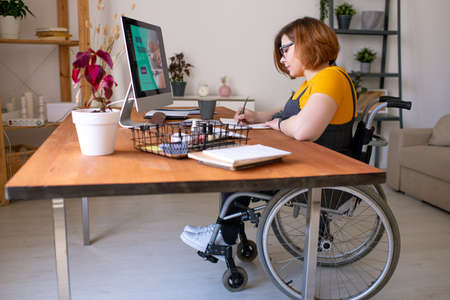Young disable woman in wheelchair making notes during remote lesson at home