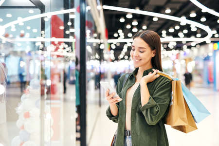 Young cheerful brunette shopper with paperbags scrolling in smartphoneの写真素材