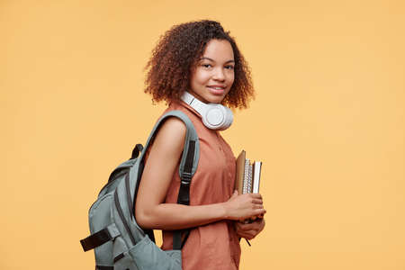 Portrait of smiling Black student girl with Afro hairstyle standing with workbooks against yellow...