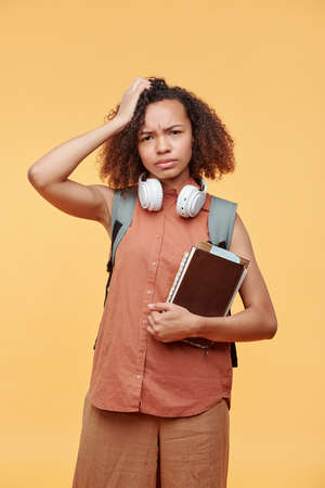 Portrait of puzzled frowning black student girl in casual outfit standing with books and scratching...