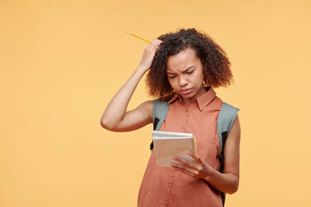 Thoughtful frowning black student girl with curly hair focused on school task reading notes in workbook against yellow backgroundの写真素材