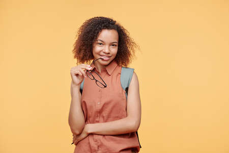 Portrait of smiling young Afro-American woman holding eyeglasses near mouth while posing against...