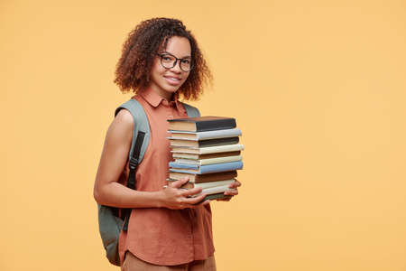 Portrait of smiling smart Afro-American student girl with satchel on back carrying stack of books against yellow backgroundの写真素材