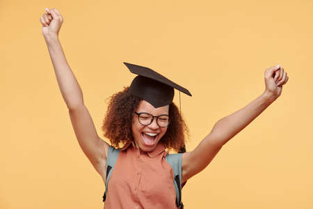 Happy excited black graduate student in eyeglasses and academic cap raising arms and screaming after completing her degreeの写真素材