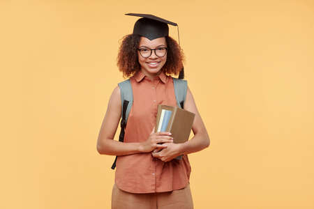 Portrait of positive successful black graduate student in cap holding stack of books against yellow...