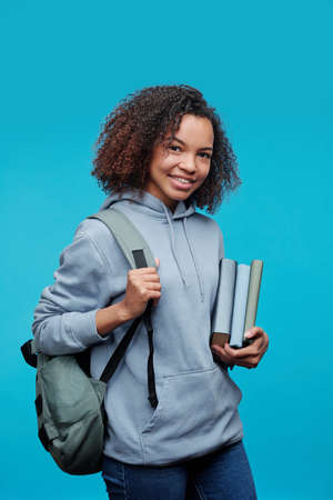 Portrait of smiling curly-haired Afro-American student girl standing with books and satchel against blue backgroundの写真素材
