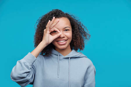 Portrait of cheerful curious black girl with Afro hairstyle looking through fingers in circle,...