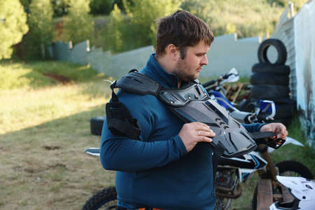 Portly young man with stubble standing outdoors and wearing motorcycle body armor while preparing...