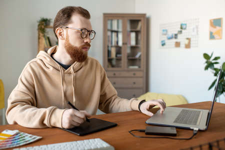 Serious creative young man in hoodie working on design project using drawing pad in home office