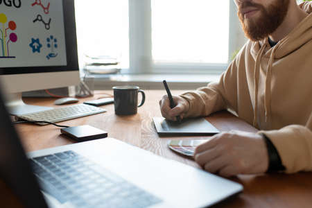 Close-up of busy brand designer sitting at desk and creating graphic design using digitizer and...