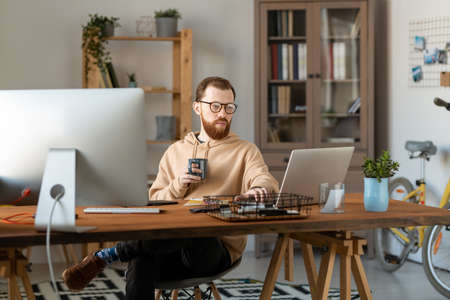 Concentrated design freelancer in eyeglasses sitting at wooden table in home office and using...