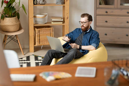 Confident young layout artist with beard sitting in yellow bean bag and using digitizer tablet while...