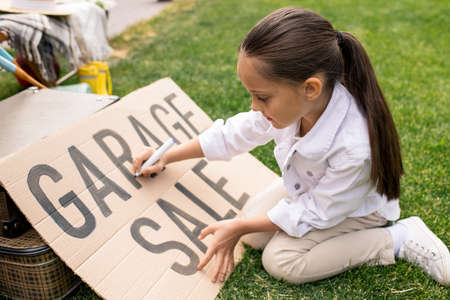 Pretty girl with ponytail sitting on grass and making banner for garage sale in backyardの写真素材