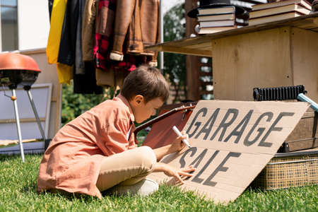 Concentrated boy in casual outfit sitting with crossed legs on ground and writing Garage Sale on carton banner