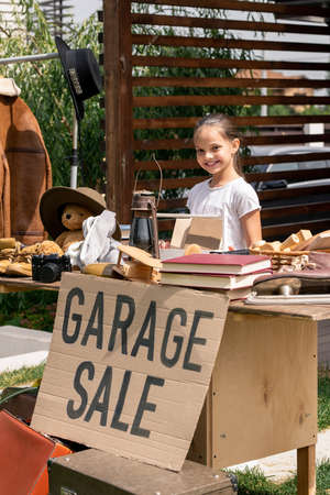 Portrait of smiling little seller standing at table with garage sale sign in backyardの写真素材