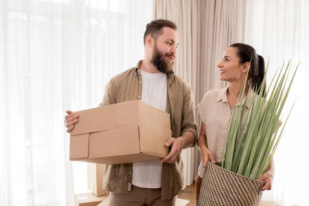 Positive young couple holding moving stuff and looking at each other while dreaming of new life in...