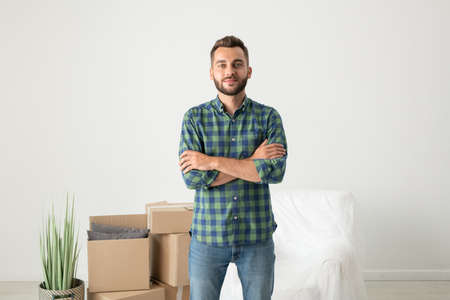 Portrait of content handsome young man in checkered shirt standing with crossed arms in new flat...