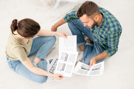 Above view of concentrated young couple sitting with crossed legs on floor and analyzing house plans...