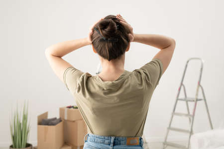 Rear view of brunette woman holding head in hands while feeling headache from fixing things in apartmentの写真素材