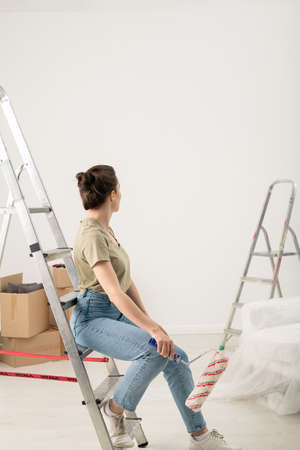 Rear view of brunette woman in jeans sitting with paint roller on step of ladder and looking at painted wallの写真素材