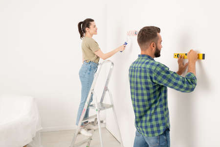 Content young woman standing on stepladder and painting wall while her boyfriend drawing line using levelの写真素材