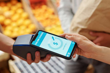 Close-up of unrecognizable woman putting smartphone to terminal while paying for products online at food marketの写真素材