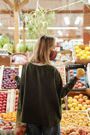 Rear view of girl in facial mask standing at fruit counter and looking at lemon while choosing it at...