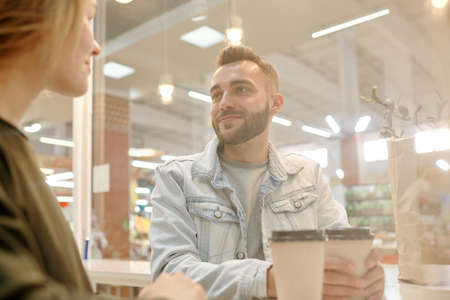 Handsome young bearded man in denim jacket sitting at counter and drinking coffee while dating with girl in fast food restaurantの写真素材