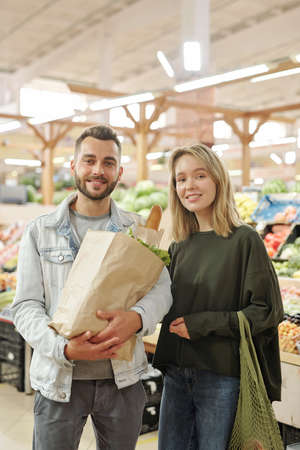Portrait of smiling beautiful young couple in casual outfits standing with bags at modern farmers...