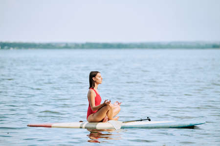 Young sportswoman in red swimsuit practicing yoga while sitting on surfboard