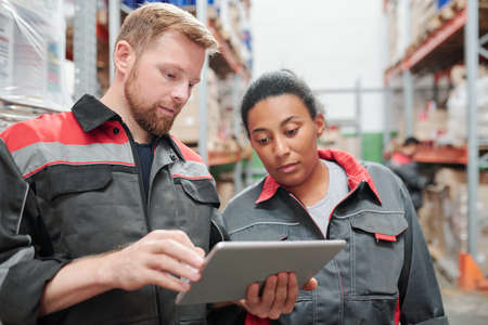 Young bearded engineer or worker of warehouse and his colleague using tabletの写真素材