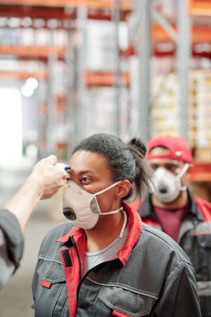 Worker of warehouse in respirator and workwear having her temperature measuredの写真素材