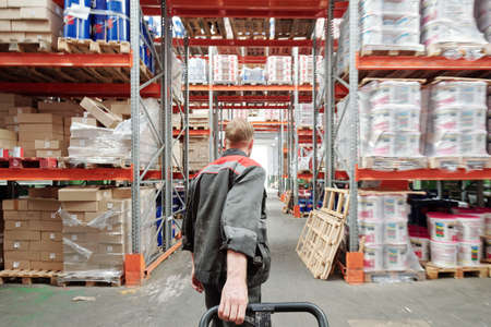 Rear view of young blond male worker of warehouse in uniform pulling loaded cartの写真素材