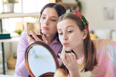 Cute teenage girl with long blond hair applying lipstick and looking in mirror