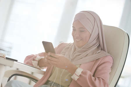 Happy young Muslim businesswoman in pink jacket and hijab sitting at table and texting sms on phone in office while listening to music in headphonesの写真素材