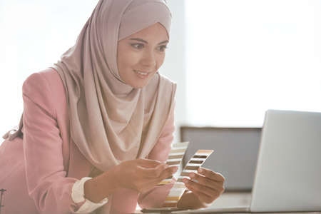 Content attractive Arabian designer in pink hijab leaning on table and comparing color palettes while talking to customer onlineの写真素材