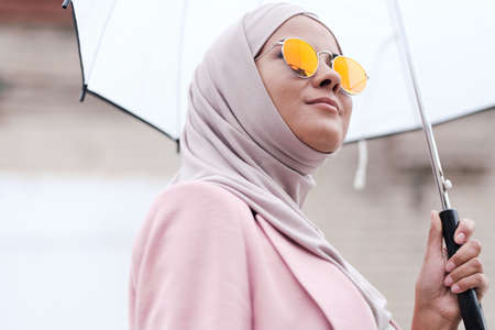 Below view of content confident young Muslim woman in sunglasses and hijab enjoying stroll under rainの写真素材