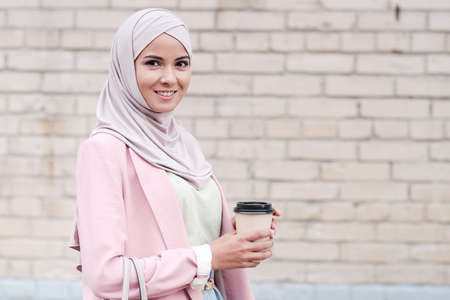 Portrait of positive beautiful young Muslim woman in hijab standing against brick wall and drinking coffee on moveの写真素材
