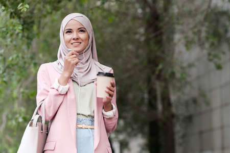 Positive confident young Muslim businesswoman in stylish outfit drinking takeaway coffee and talking through phone headset outdoorsの写真素材