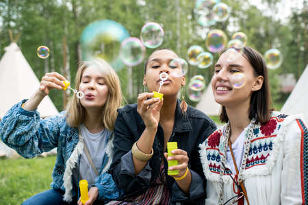 Carefree young multi-ethnic women blowing soap bubbles while relaxing at forest campsite