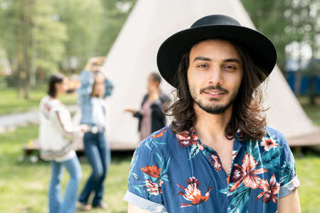 Portrait of content handsome Jewish guy in black hat and shirt with flower print resting with friends at forest campsiteの写真素材