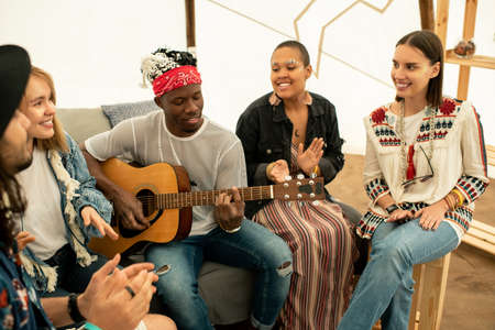Positive young black musician playing guitar and singing song with friends in tent