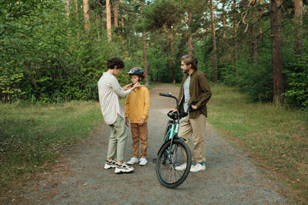 Young careful mother helping her son with tying up belts of sports helmet
