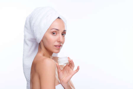 Young gorgeous woman with white towel on head holding jar of facial creamの写真素材