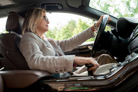Side view of mature woman sitting by open window in car and driving to work