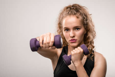 Pretty young sportswoman with blond curly hair stretching arm with dumbbellの写真素材