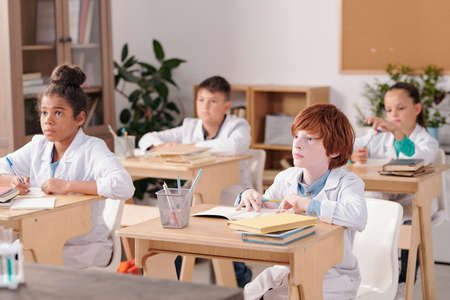 Group of intercultural elementary schoolkids in whitecoats sitting by desksの写真素材