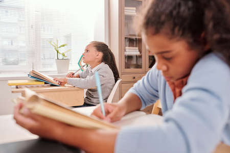 Pensive schoolgirl with open book thinking of answer to question on blackboardの写真素材